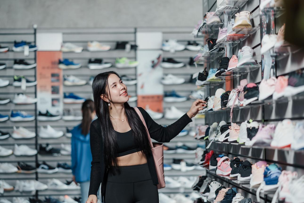 Asian woman browsing sneakers display in modern sport shop. Shopping for sports shoes, fitness fashion, activewear, consumer behavior, retail shopping experience.